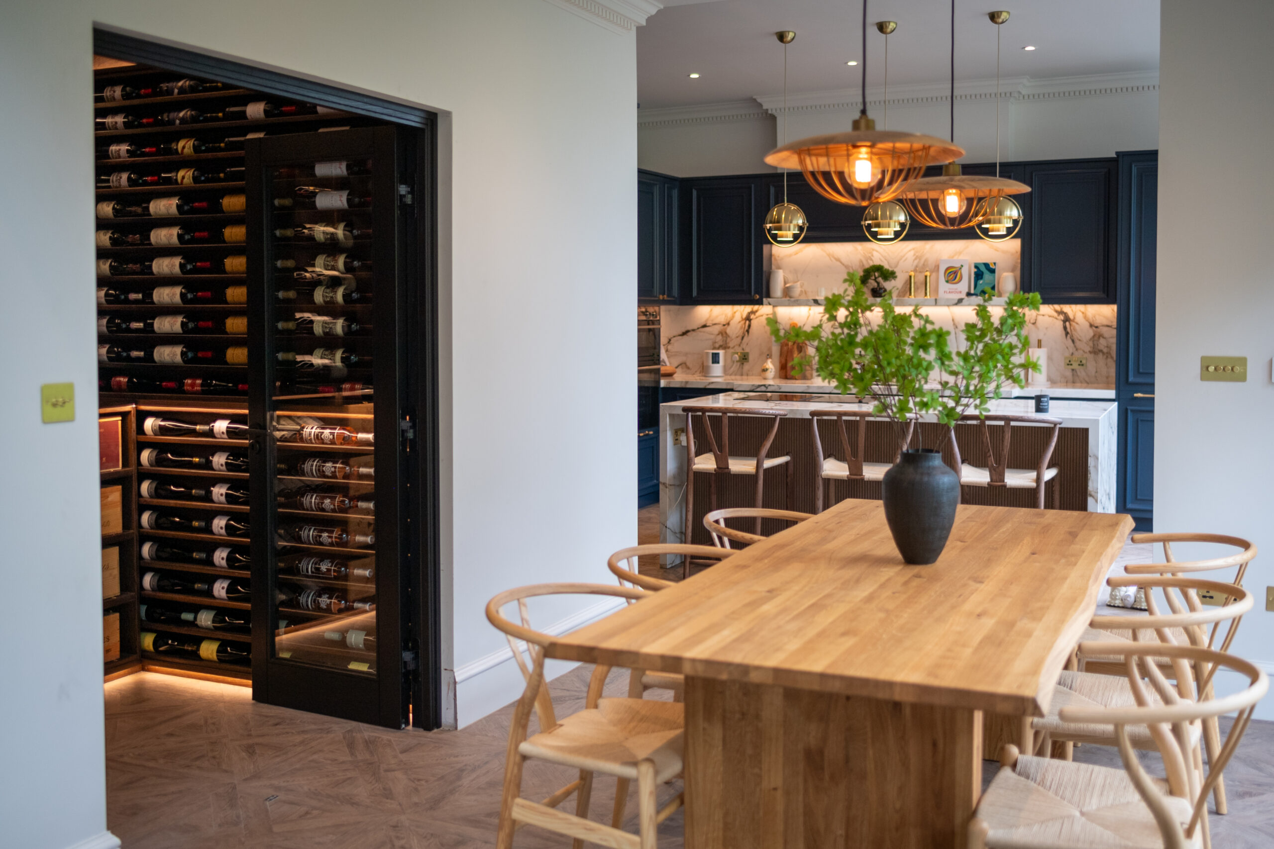 A Sorrells wine cellar, integrated into a traditional style kitchen. The wine cellar features dark oak racking, whilst the kitchen has a light oak table and blue cabinets with a marble work bench.